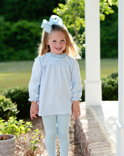 Young girl in a light blue dress with a bow in her hair, standing outdoors.
