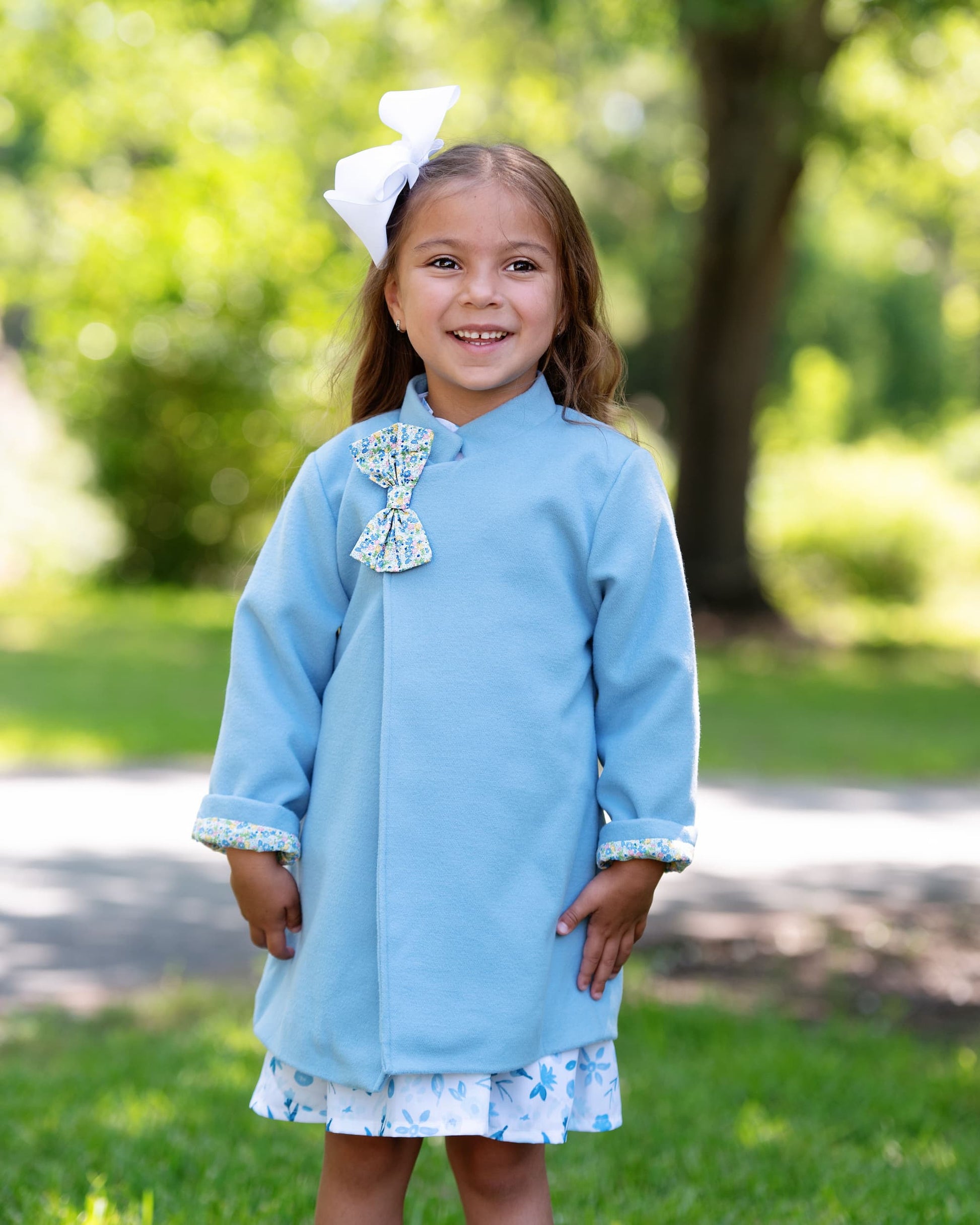 Young girl in a light blue dress with a large bow standing outdoors.