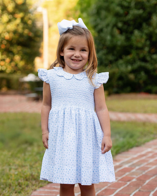 Young girl in a light blue dress with a white bow in her hair, standing outdoors.