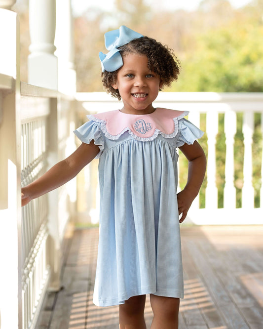 Young girl in a light blue dress with pink collar standing on a porch.