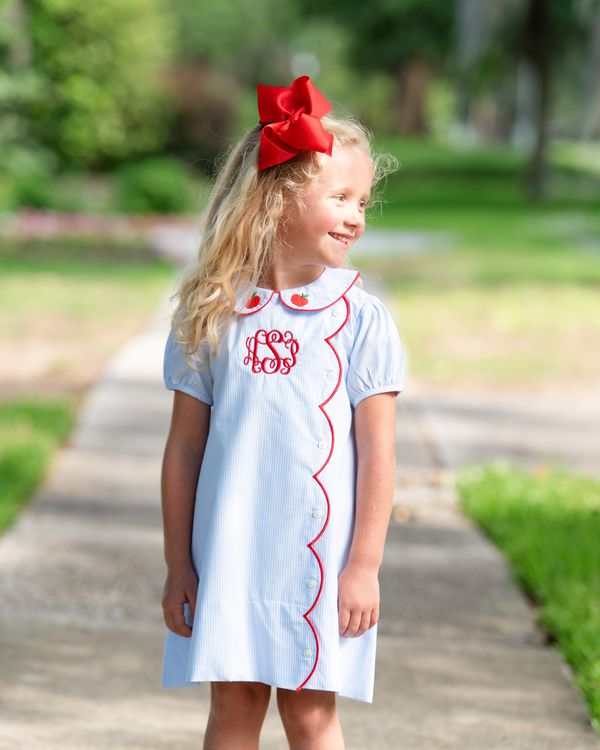 Young girl in a light blue dress with red trim and bow standing outdoors.