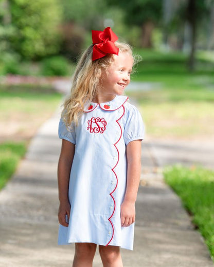 Young girl in a light blue dress with red trim and bow standing outdoors.
