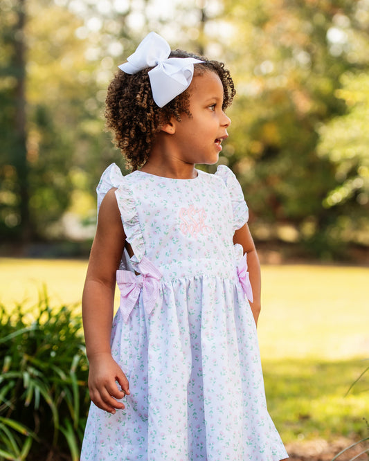 Young girl in a light floral dress with pink bows and a large white bow in her hair , standing outdoors.