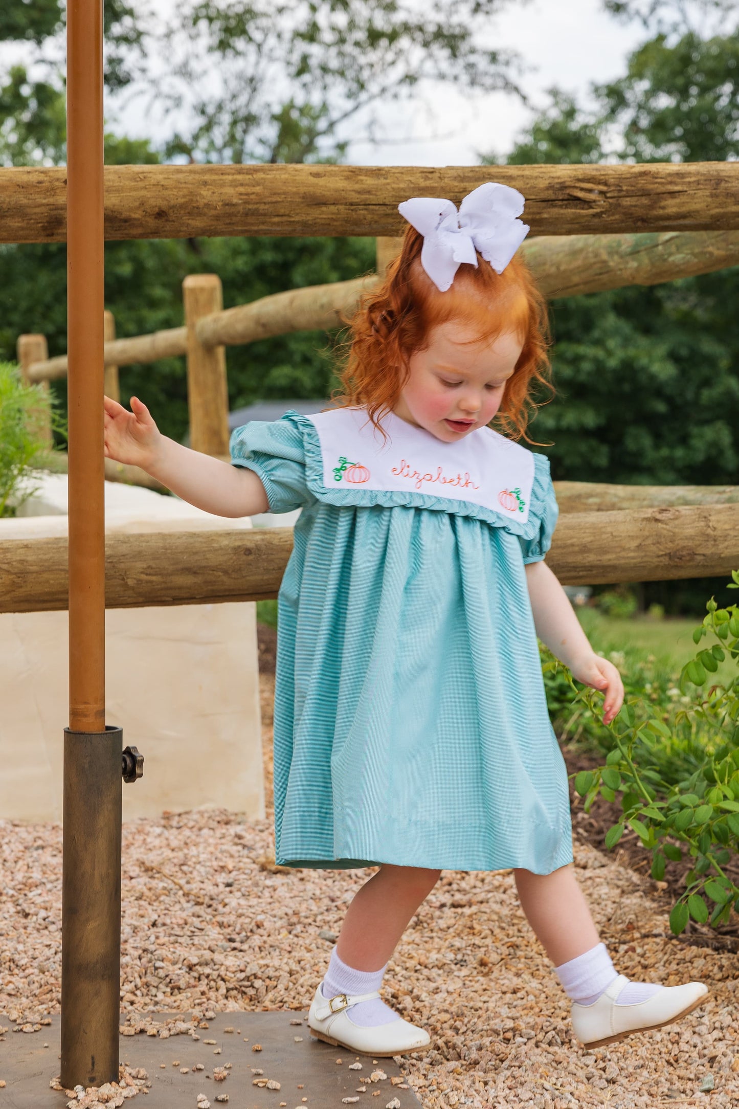 Young girl in a light green dress with a white bow standing outdoors near a wooden fence.