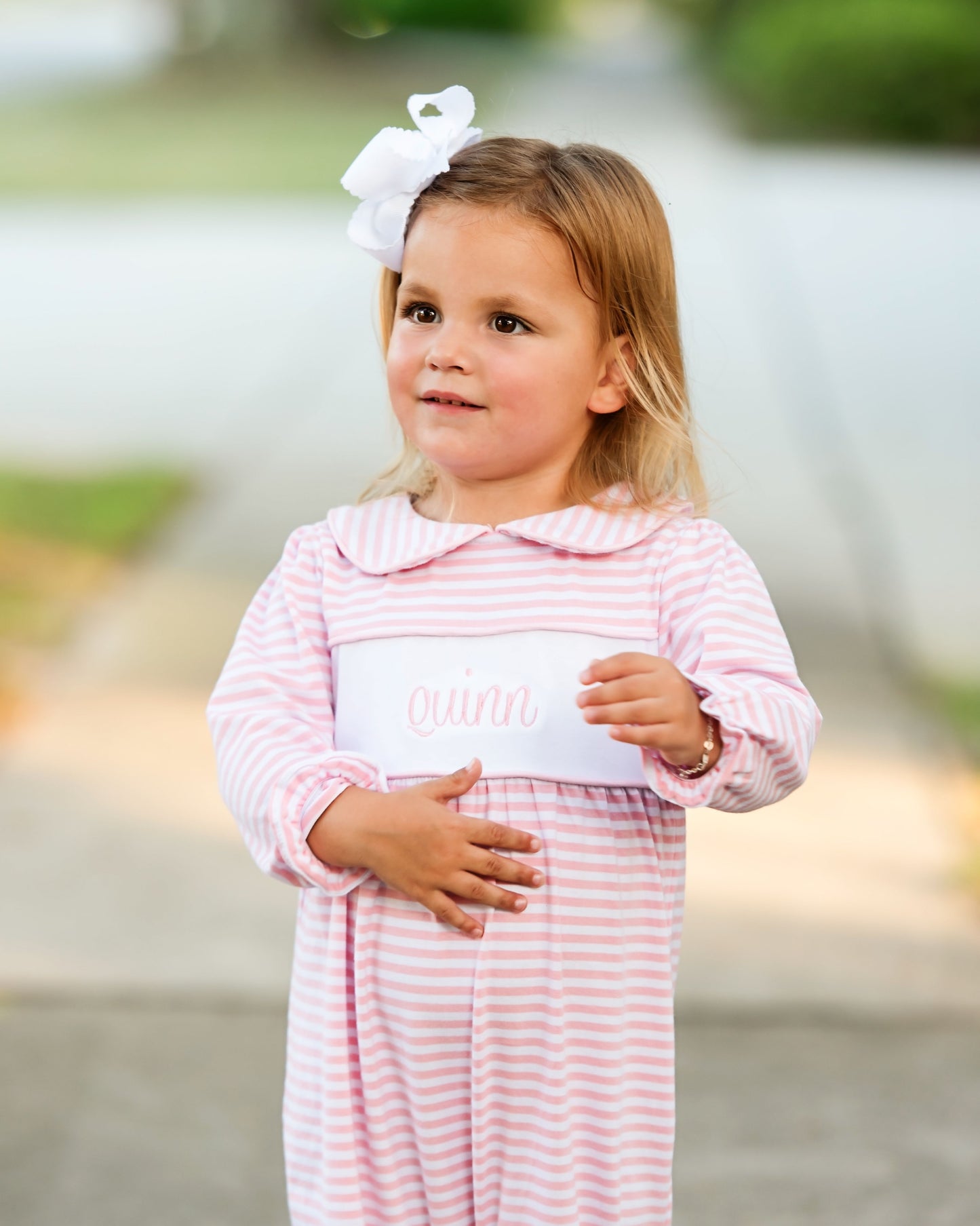 Young girl in a pink and white striped dress with a white bow in her hair, standing outdoors.