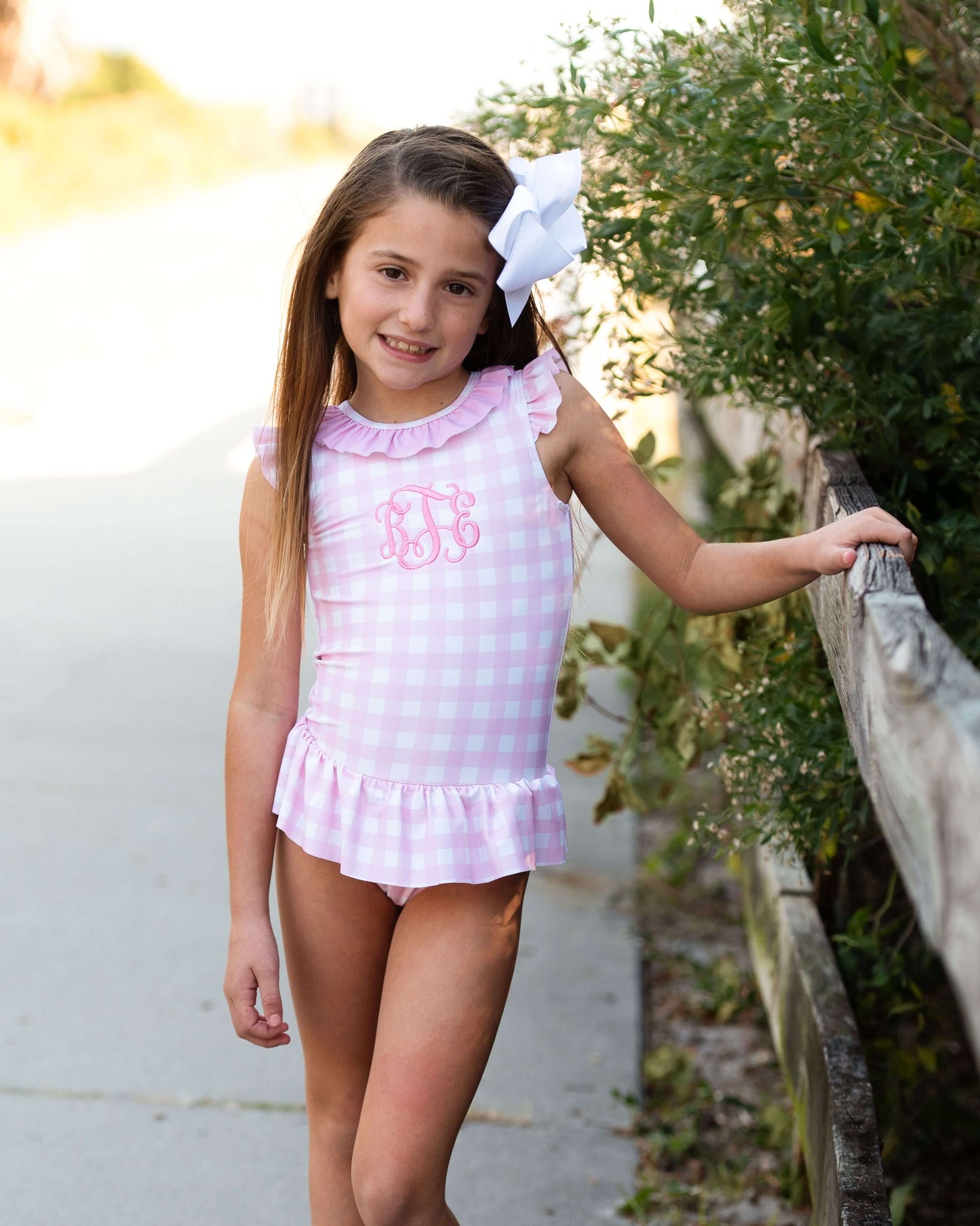 Young girl in a pink checkered dress standing outdoors with greenery in the background