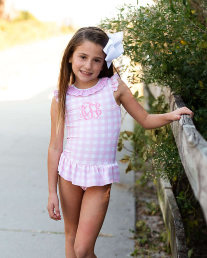 Young girl in a pink checkered dress standing outdoors with greenery in the background