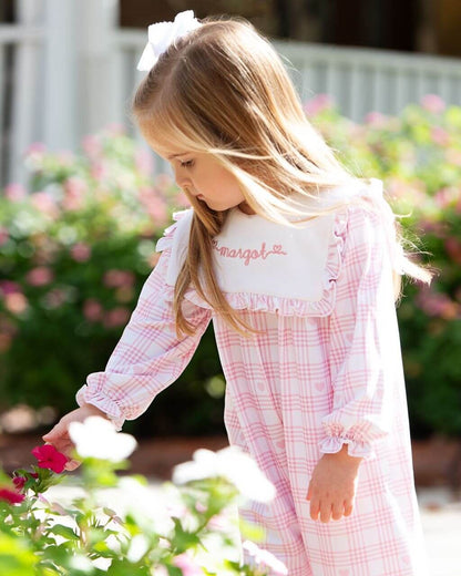 Young girl in a pink checkered dress with a white bow in her hair, standing in a garden.