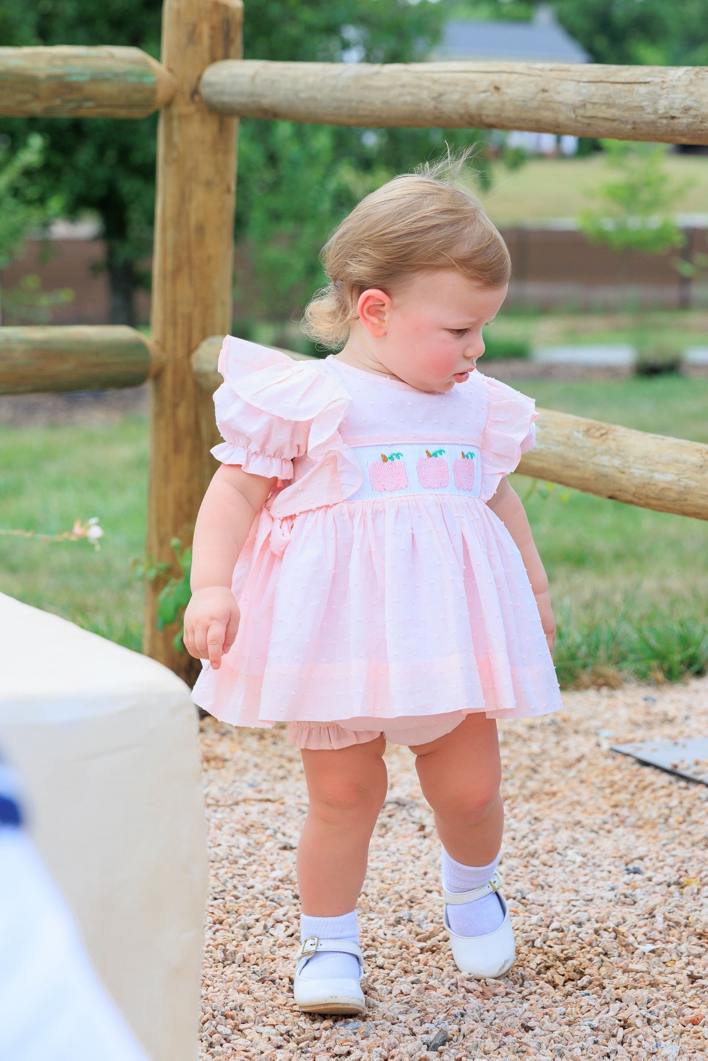 Young girl in a pink diaper set standing near a wooden fence outdoors