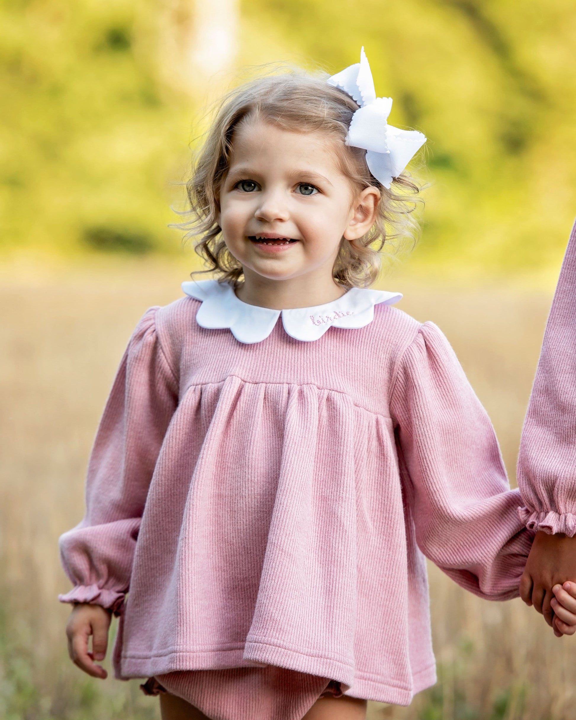 Young girl in a pink diaper set with a white collar and bow in her hair, standing in a field.