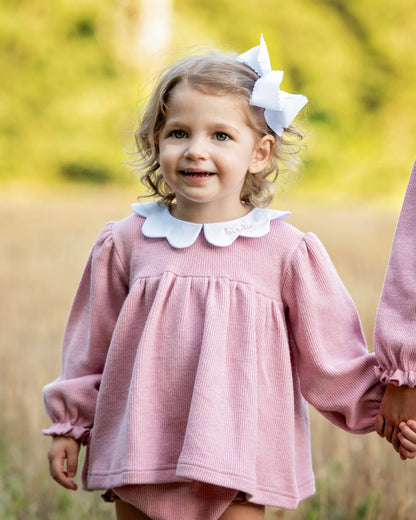Young girl in a pink diaper set with a white collar and bow in her hair, standing in a field.