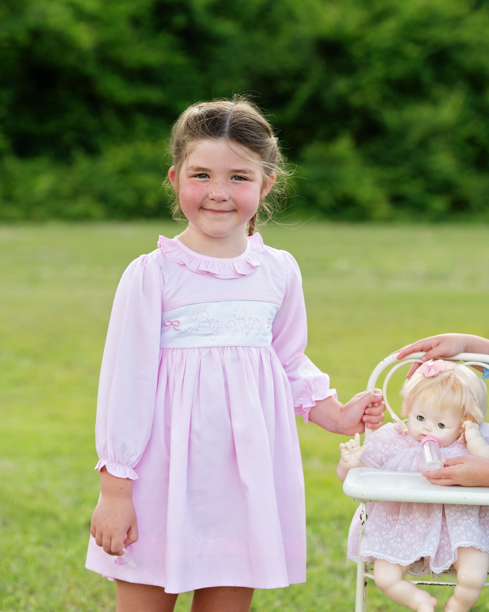 Young girl in a pink dress holding a doll in a grassy outdoor setting