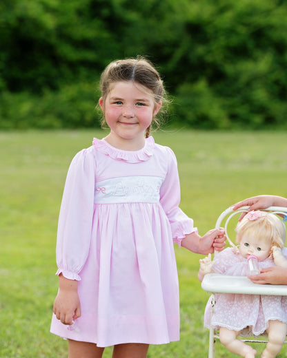 Young girl in a pink dress holding a doll in a grassy outdoor setting