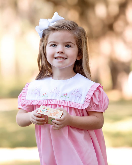 Young girl in a pink dress holding blocks outdoors with a blurred natural background