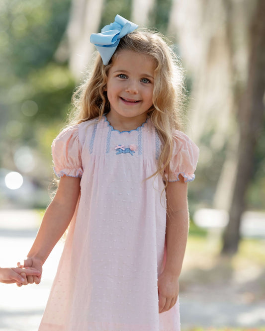 Young girl in a pink dress with a blue bow in her hair, standing outdoors.
