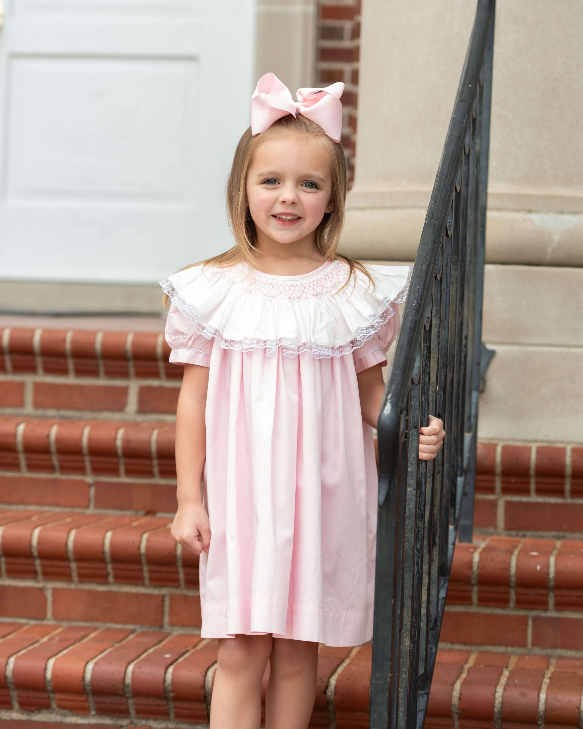 Young girl in a pink dress with a large bow standing on a staircase.