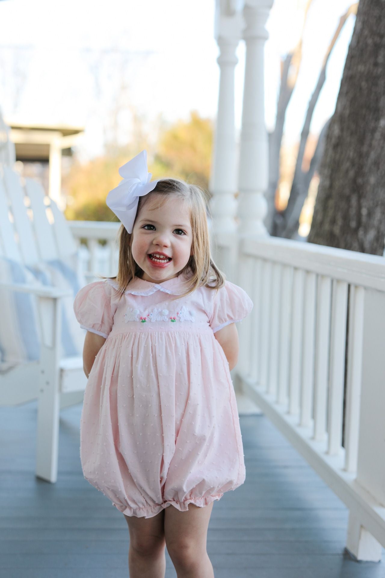 Young girl in a pink dress with a large white bow standing on a porch.