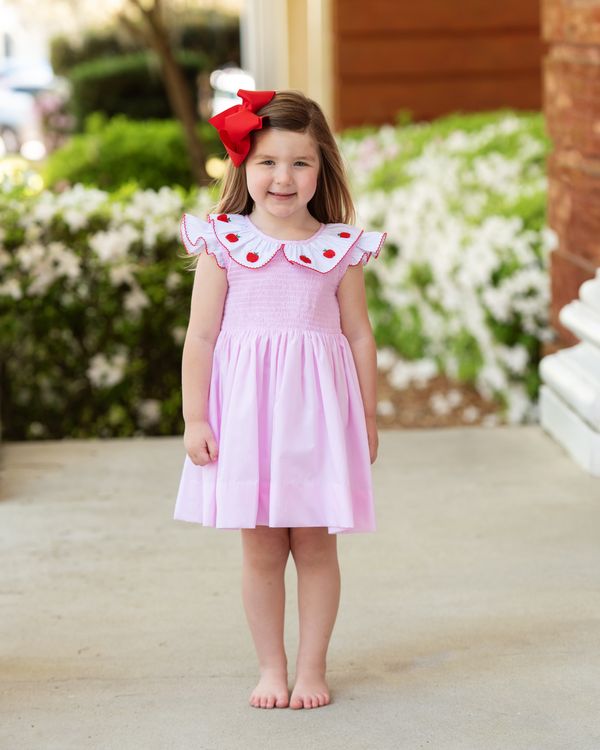 Young girl in a pink dress with a red bow standing outdoors.