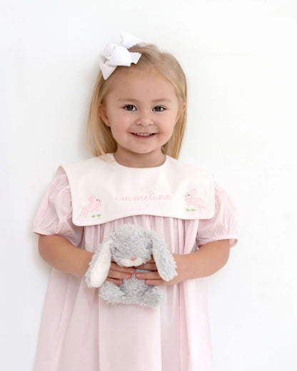Young girl in a pink dress with a white bib holding a gray teddy bear against a white background