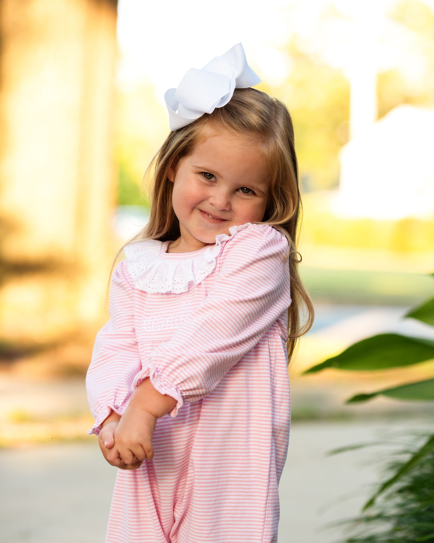 Young girl in a pink dress with a white bow in her hair, standing outdoors with a blurred natural background.