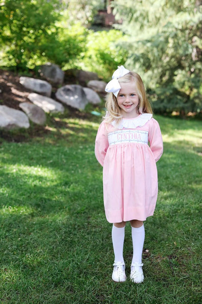 Young girl in a pink dress with a white bow standing on grass with trees in the background