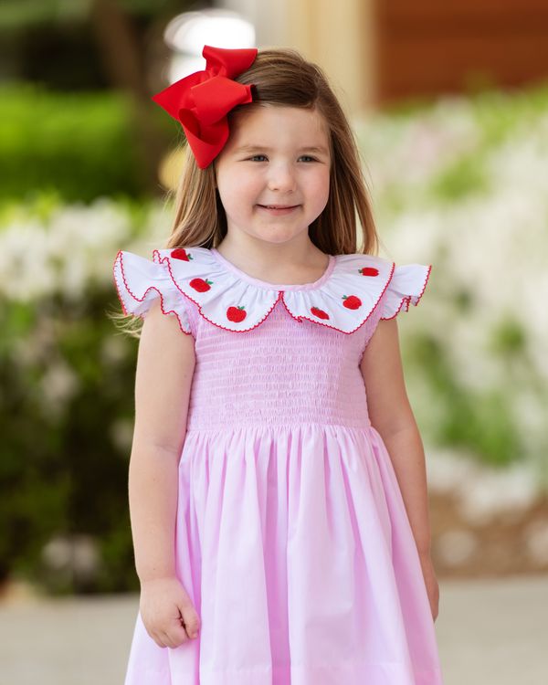 Young girl in a pink dress with red heart details, smiling outdoors.