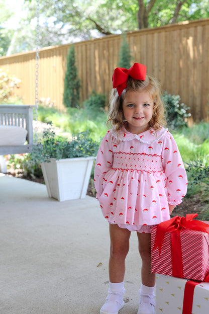 Young girl in a pink dress with red polka dots and a large red bow, standing outdoors next to a gift box.