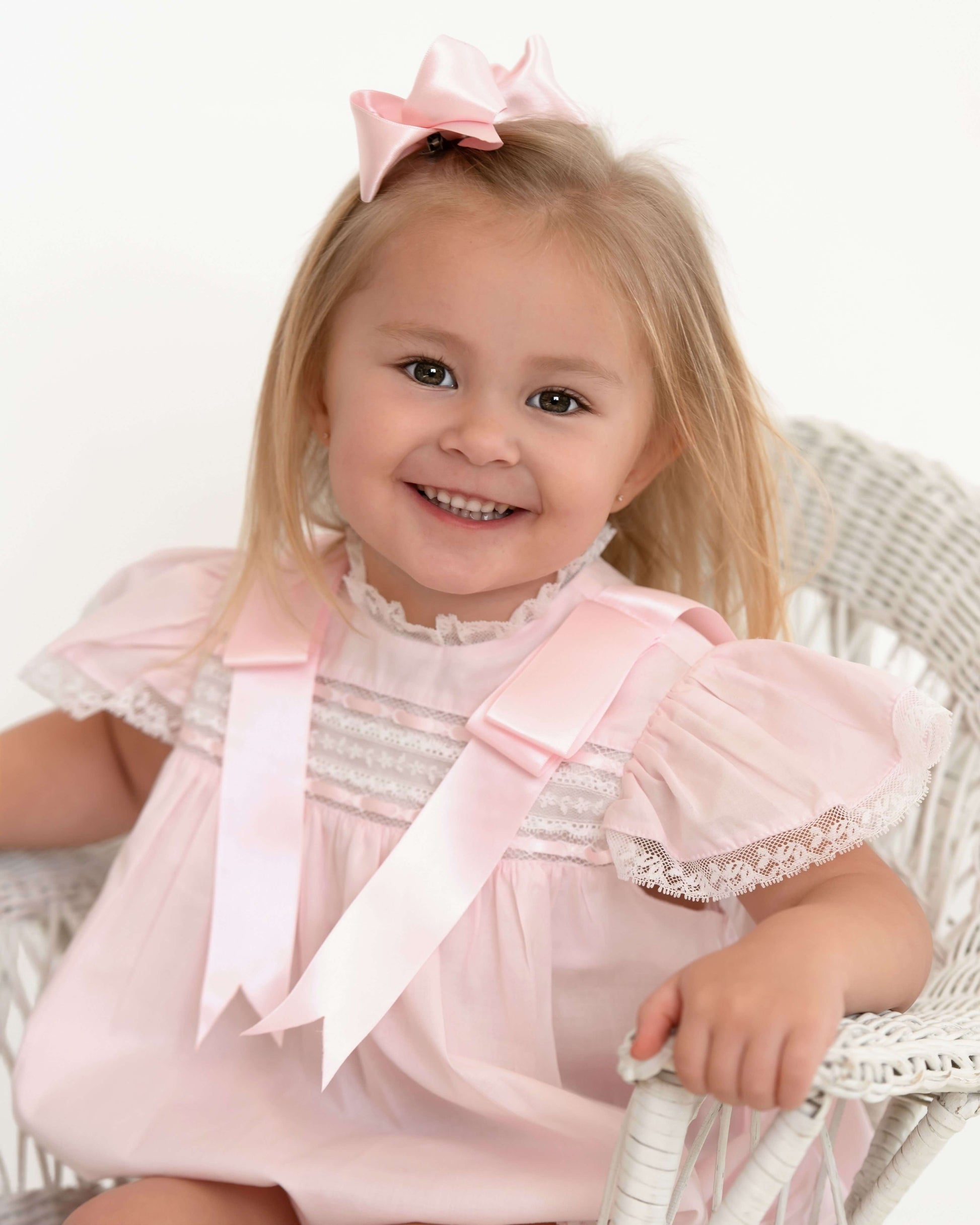 Young girl in a pink dress with ruffles and a bow, sitting on a white chair.
