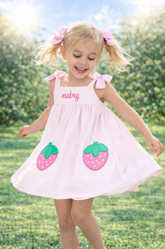 Young girl in a pink dress with strawberry designs and 'mabry' embroidered on it, standing in a grassy field.
