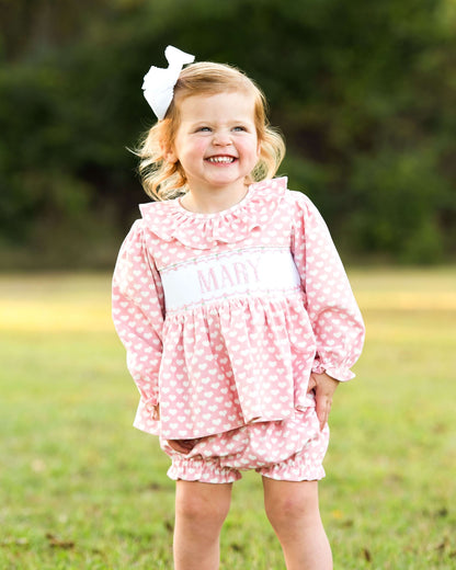 Young girl in a pink heart outfit with a large bow in her hair, standing outdoors.