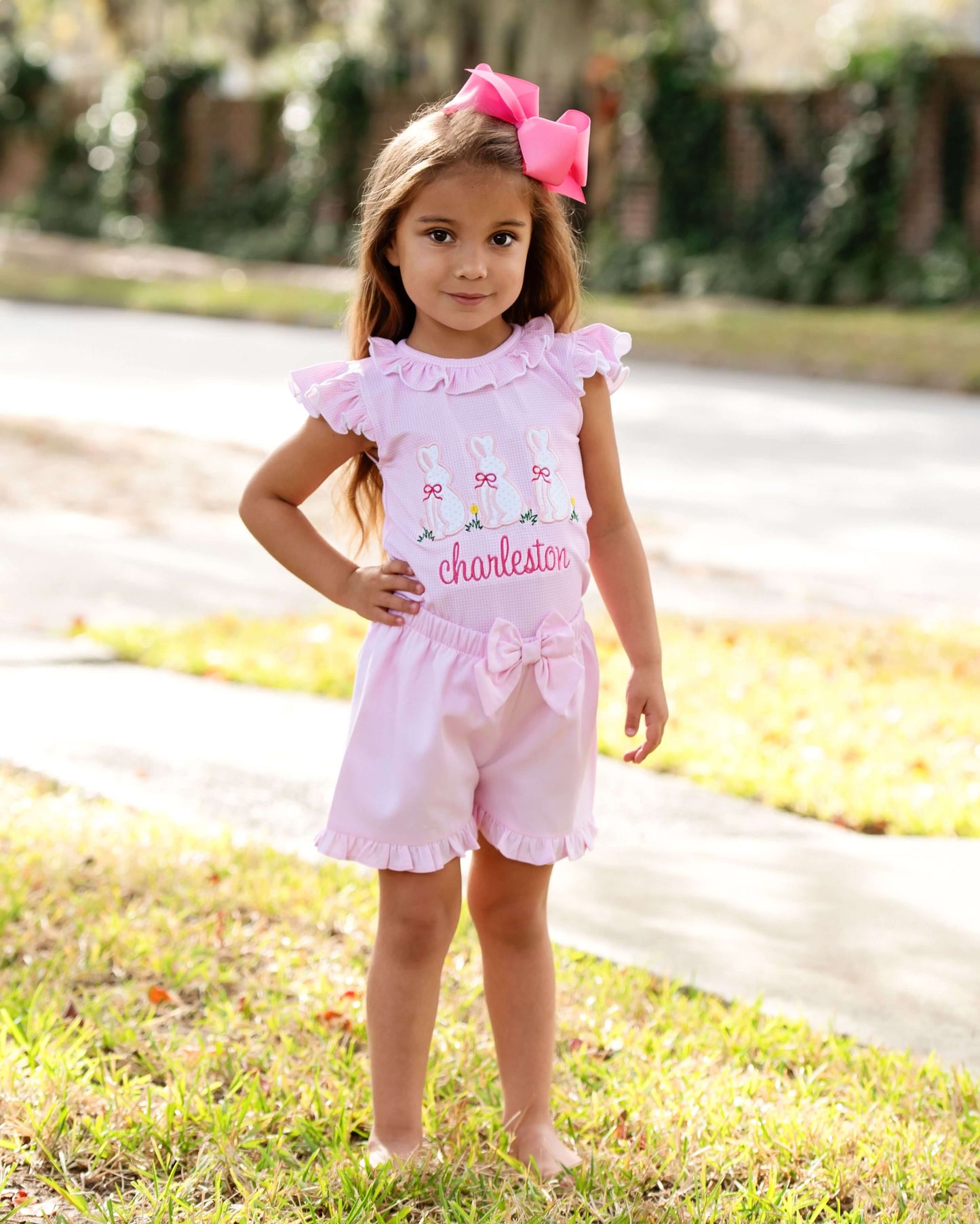 Young girl in a pink outfit with a bow standing on grass.
