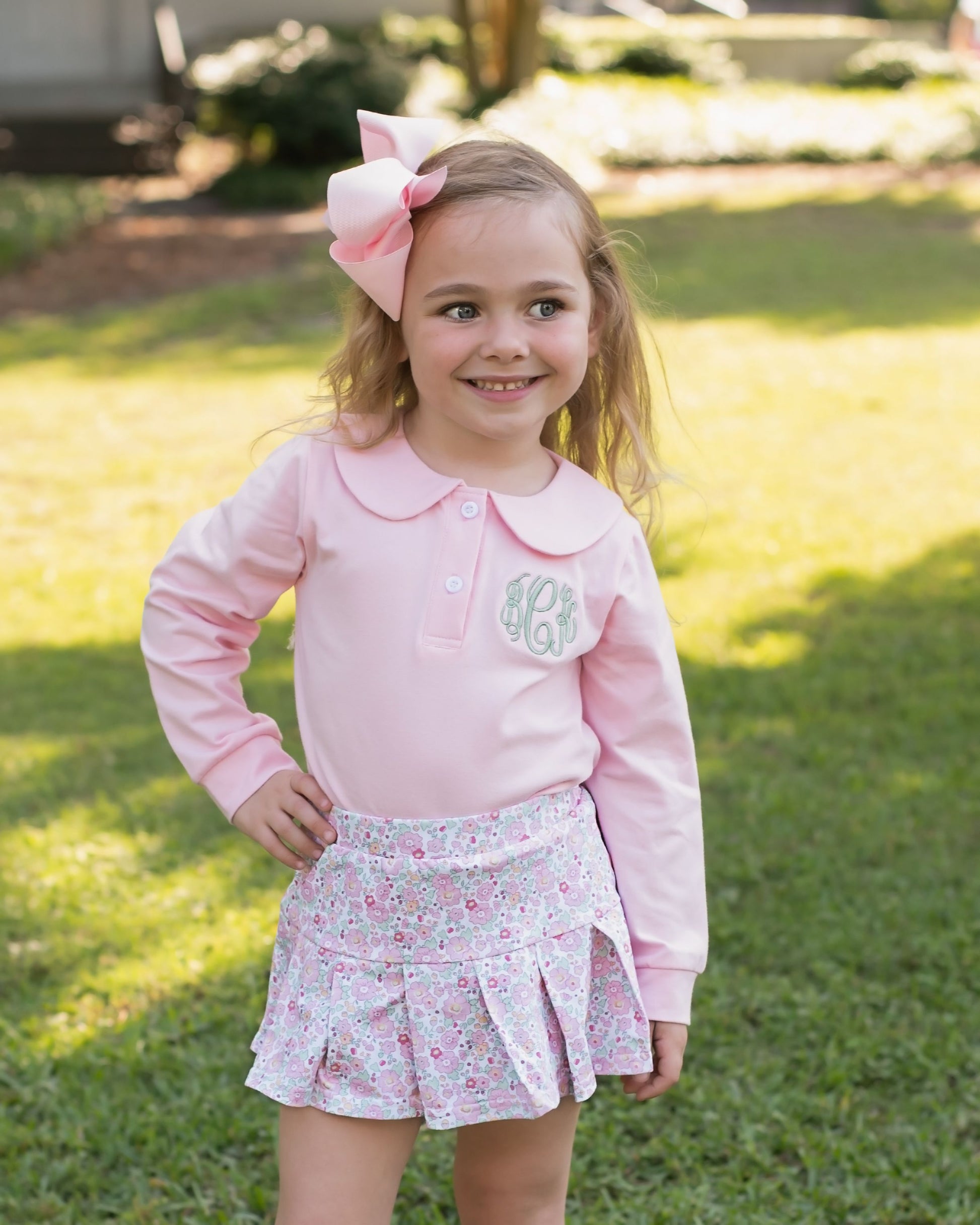 Young girl in a pink outfit with a floral skirt and large bow outdoors.
