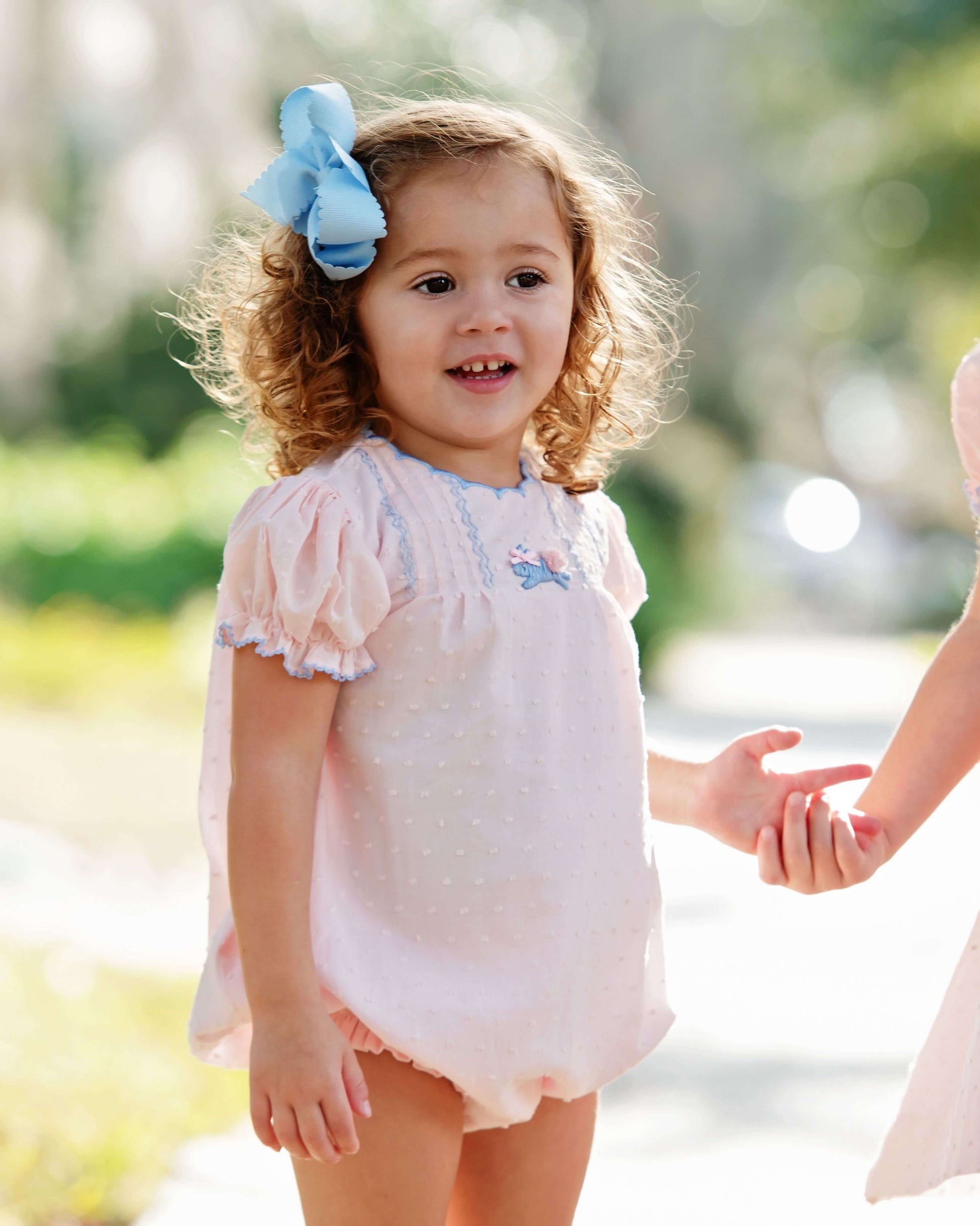 Young girl in a pink swiss dot outfit with a blue bow, standing outdoors.