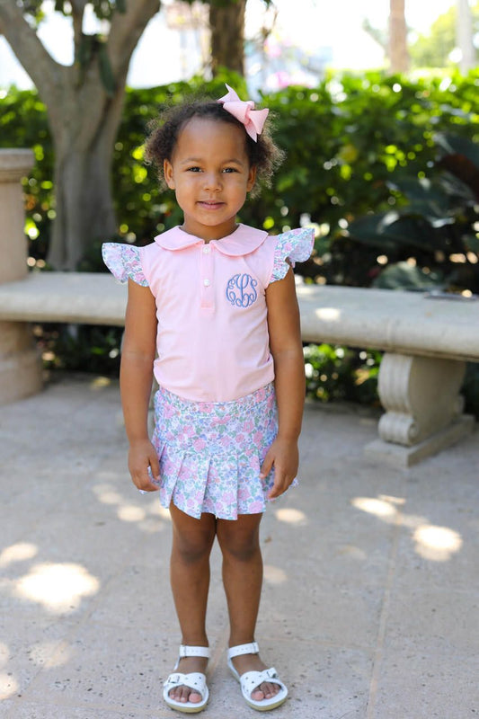 Young girl in a pink top and floral skirt standing outdoors with greenery in the background