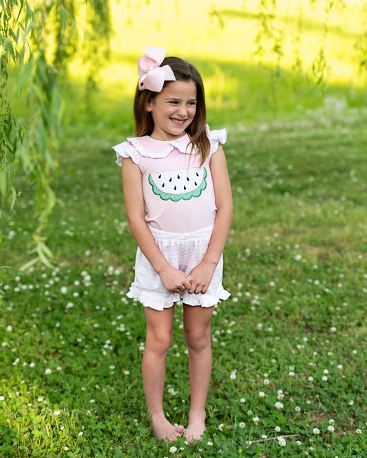 Young girl in a pink top with watermelon design and white shorts standing in a grassy field.