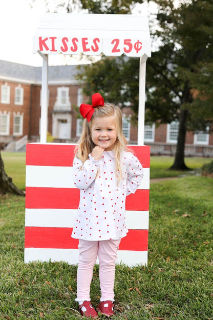 Young girl in a polka dot dress standing in front of a 'Kisses 25 cents' sign outdoors.