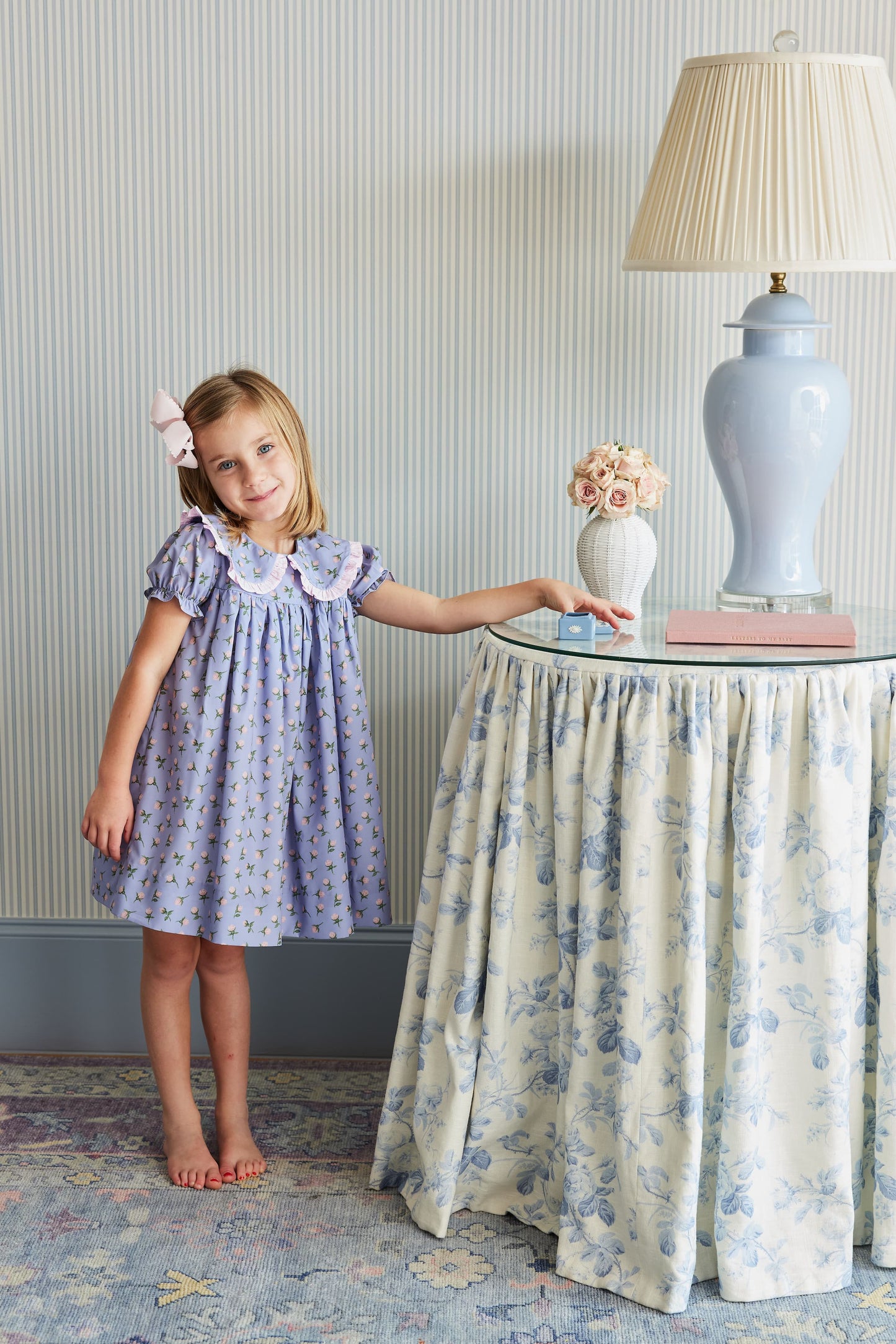 Young girl in a purple dress standing next to a table with decorative items in a room.