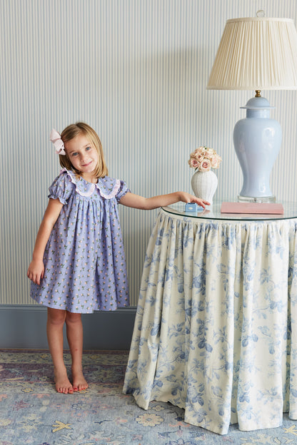 Young girl in a purple dress standing next to a table with decorative items in a room.