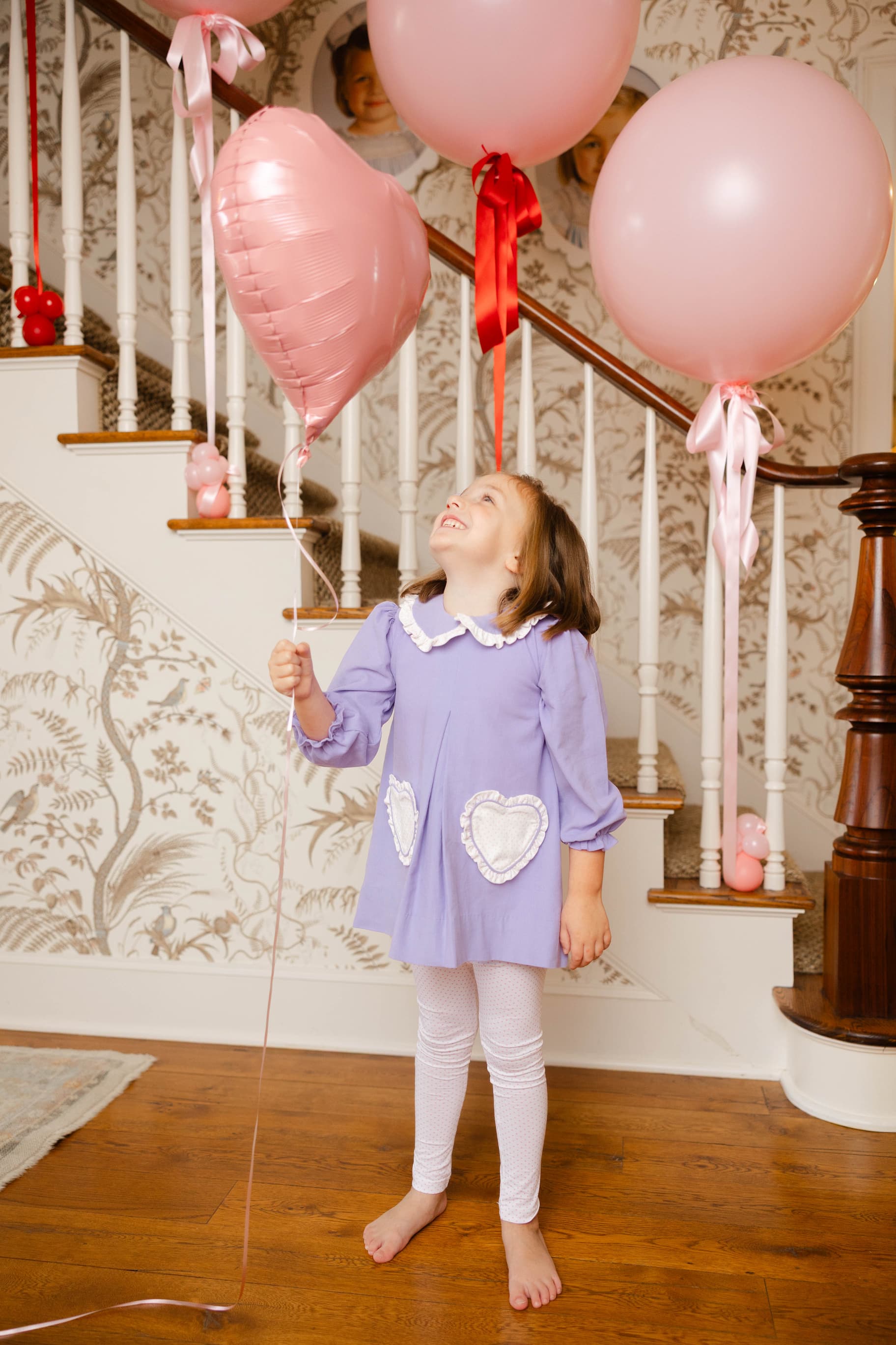 Young girl in a purple dress standing on a wooden floor with pink balloons tied to a staircase.