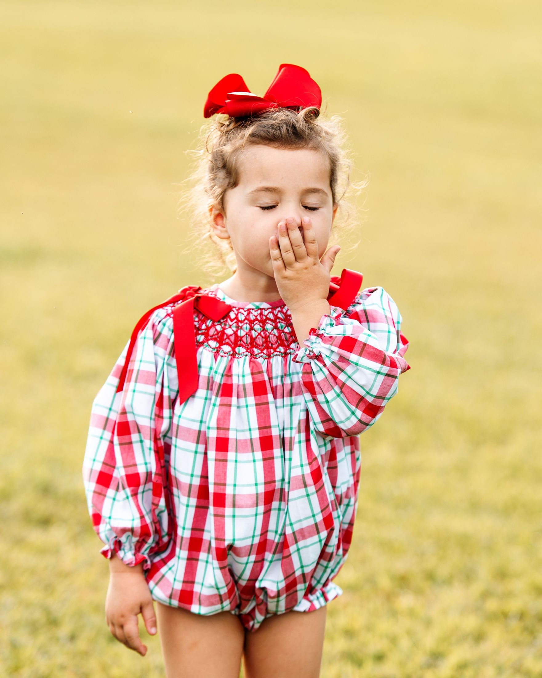 Young girl in a red and white checkered outfit with a red bow in her hair, standing in a field.