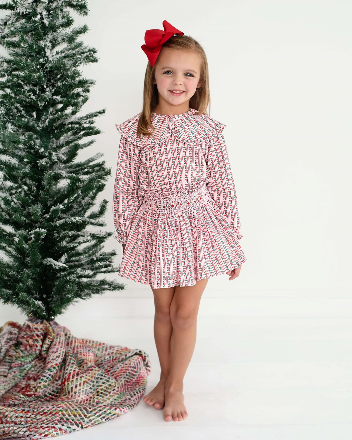 Young girl in a red and white floral skirt set with a red bow standing next to a  Christmas tree.