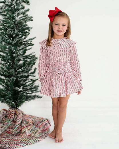 Young girl in a red and white floral skirt set with a red bow standing next to a  Christmas tree.