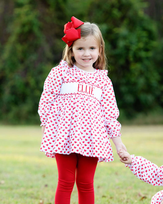 Young girl in a red and white heart legging set with a large red bow outdoors.