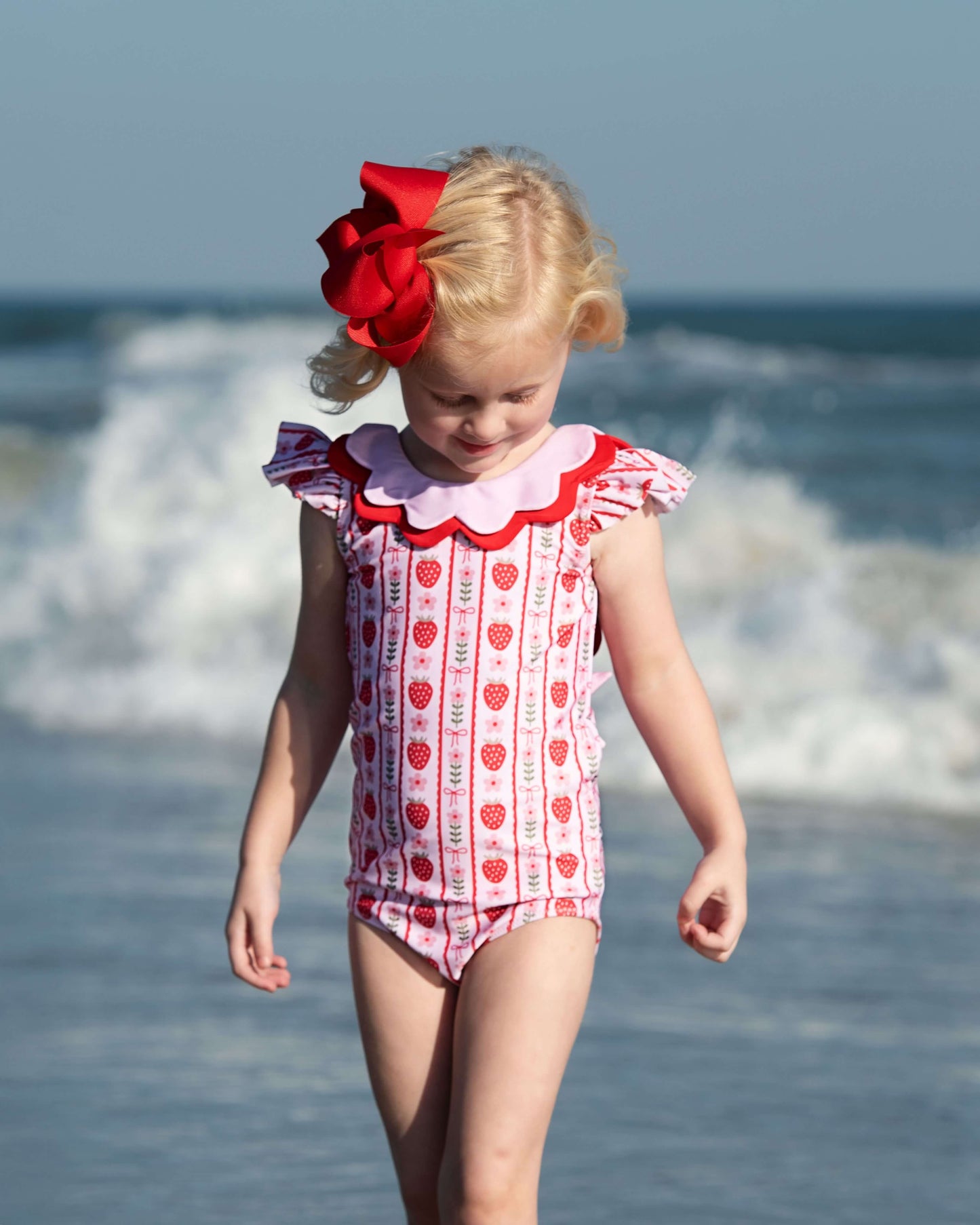 Young girl in a red and white swimsuit with a large red bow on her head, standing on a beach.