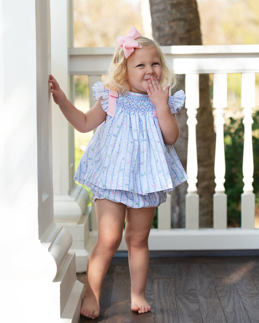 Young girl in a smocked diaper set with pink bows, standing on a wooden deck.