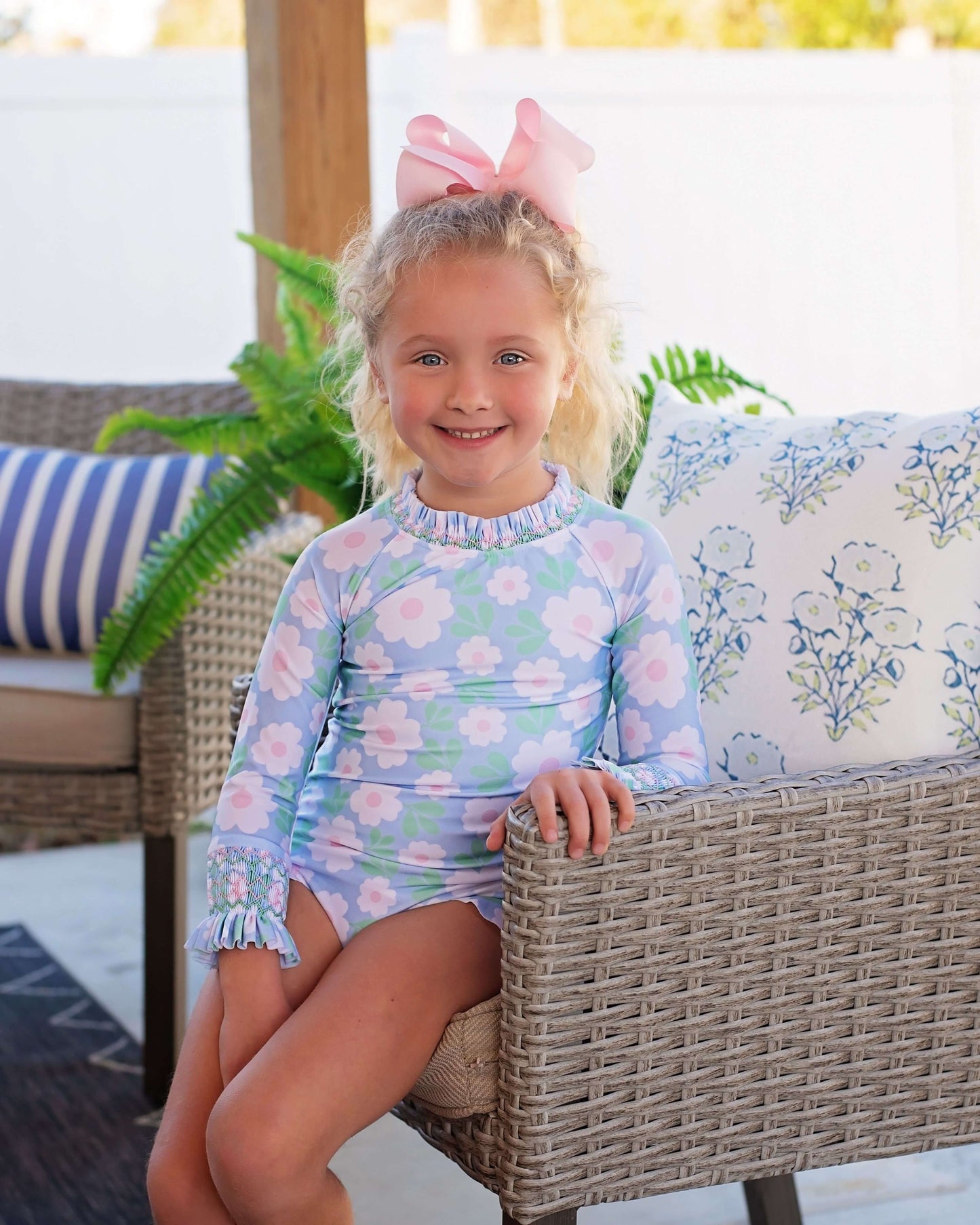 Young girl in a smocked floral swimsuit sitting on a wicker chair outdoors.