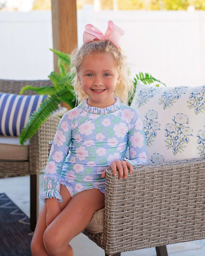 Young girl in a smocked floral swimsuit sitting on a wicker chair outdoors.