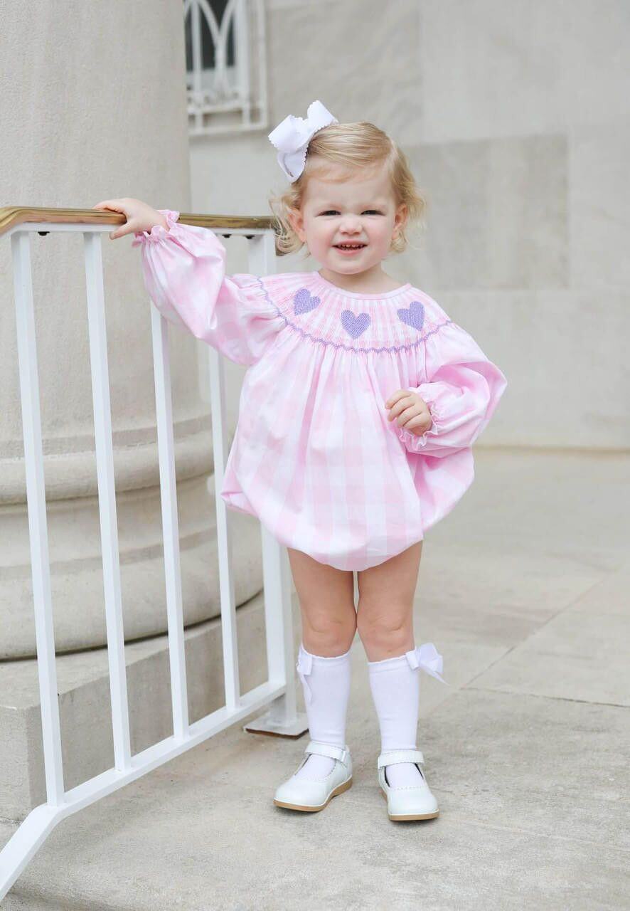 Young girl in a smocked pink check bubble with heart patterns standing next to a white railing.