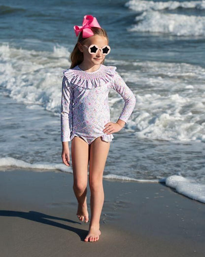 Young girl in a swimsuit and sunglasses standing on a beach with ocean waves in the background