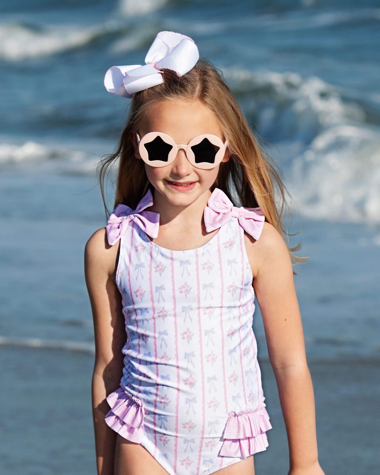 Young girl in a swimsuit with a bow and sunglasses standing by the water.